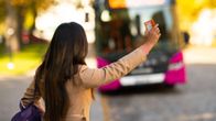 En person håller upp ett busskort mot en ankommande buss med oskarp skylt i en stadsmiljö med höstträd.
