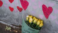 Kimppu keltaisia ruusuja nojallaan muuria vasten. Muuriin on maalattu  punaisia sydämiä, joissa on koronapandemiaan kuolleiden nimiä. Kukkakimppua ympäröivään muoviin on kirjoitettu syntymäpäiväonnittelu vainajalle.