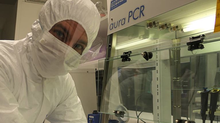 Valkoiseen huppuhaalariin, sujalaseihin ja kumihanskoihin pukeutunut mies laboratoriapöydän ääressä. Hänen kädessään on pieni muovipussi ja sen sisällä luunpalanen.