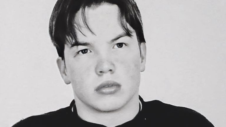 En svartvit bild på Janne Raninen tagen hos polisen. Han har en svart t-skjorta på sig och på bröstkorgen står en vit lapp med fångnummer och identifikationsnummer.