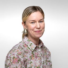 Nainen katsoo kameraan vaaleanpunaisessa paidassa.
