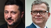 Kaksi pukuihin pukeutunutta henkilöä seisoo vierekkäin; taustalla vaaleat seinät.
