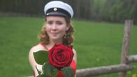 Anna Pursiainen ojentaa ruusua kohti kameraa päässään ylioppilaslakki ja taustallaan niitty.