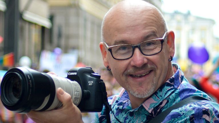 Mies katsoo kameraan, käsissään kamera isolla teleobjektiivilla. Kuvan päällä teksti "Osaamistani olen päässyt hyödyntämään usein ja monipuolisesti, kuvaamisen ja leikkaamisen ohessa myös tulkkina ja kääntäjänä sekä soittajana radio- ja tv-ohjelmissa" Berislav, kuvaaja-leikkaaja