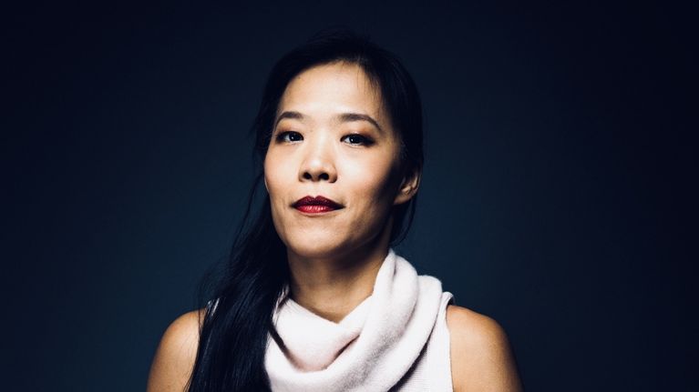 Taiwanilainen nainen vaaleassa hihattomassa neuleessa istuu tummansinistä taustaa vasten.