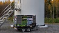 Dieselgenerator står på hjul nedanför ett vindkraftverk. Det går en sladd från generatorn in i kraftverket.