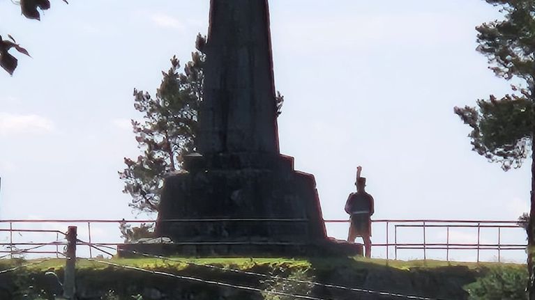 Ett monument i sten står på en höjd med räcke, en person syns intill monumentet, träd och elledningar i förgrunden.