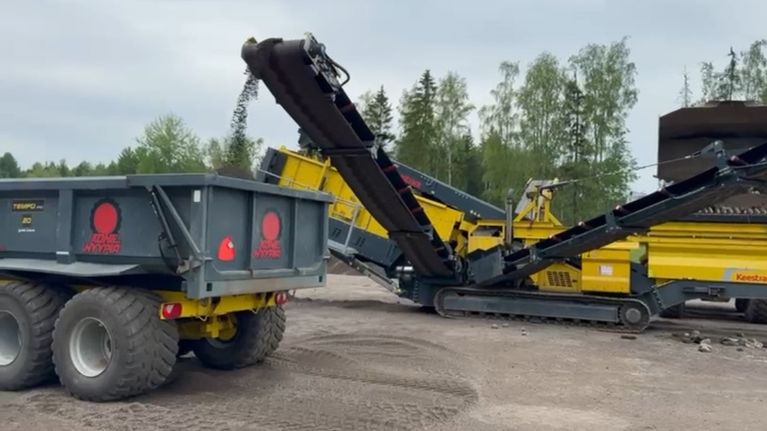 Kentuck-merkkinen seulontakone seuloo maa-ainesta ja syöttää sitä maansiirtoajoneuvon kippilavalle. Taustalla näkyy metsää ja pilvinen taivas.