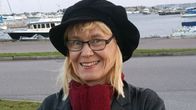 Silmälasipäinen vaaleatukkainen nainen hymyilee. Hänellä on päässään iso musta lippalakki ja kaulassa punainen huivi. Hänen takanaan näkyy venesatama.