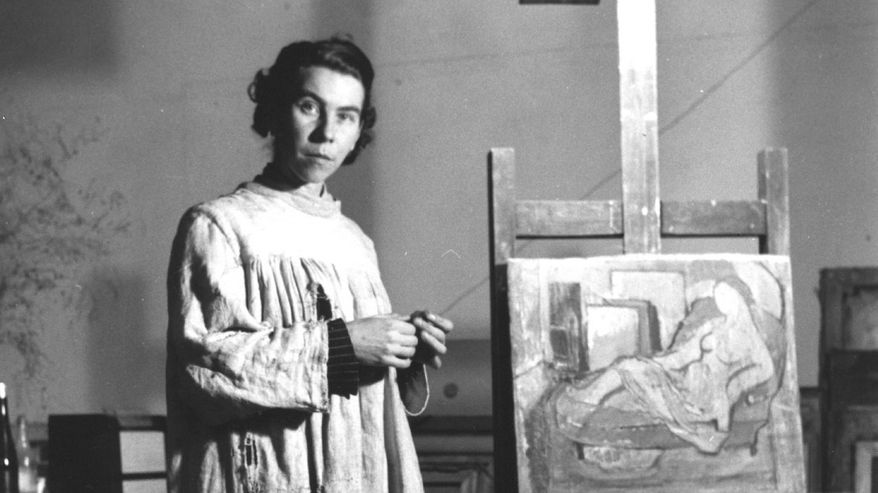 Taiteilija Tove Jansson seisoo maalaustelineen luona päällään vaalea taidemaalarin työtakki.