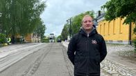 Mies seisoo kivituhkapäällysteisellä kadulla, jonka varrella näkyy keltaisia taloja ja vehreitä puita. Taustalla on parkkeerattu kuorma-auto.