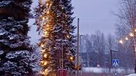 Jouluvalot saanut, kapea ja pitkä joulukuusi lumisessa kylämaisemassa. 