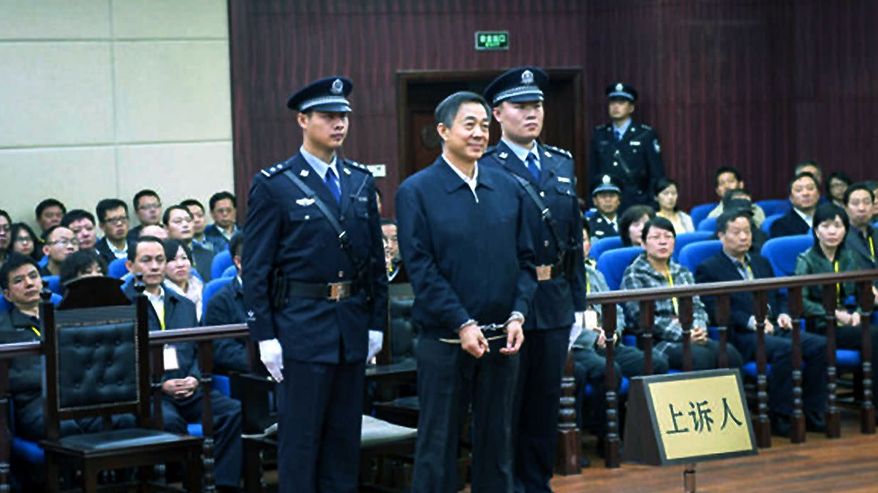 Bo Xilai seisoo käsiraudoissa oikeudessa, ympärillä kaksi poliisia univormussa. Bolla on hieman pilkallinen hymy. 
