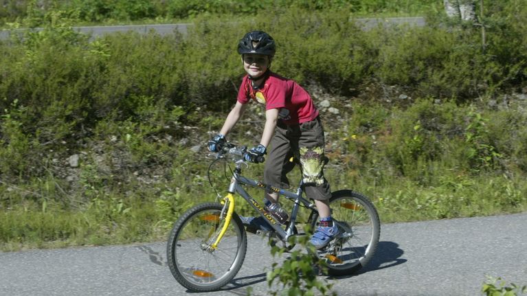 En liten pojke iklädd röd t-tröja och cykelhjälm cyklar med en mountainbike på en landsväg. 