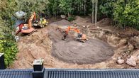 Bild tagen uppifrån, kanske med drönare. Nere till vänster syns taket på ett hus, framför det en stor grop. I gropen jobbar en orange grävskopa med att breda ut grus, invid gropen står en annan och en traktor.