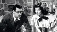 Cary Grant ja Katharine Hepburn elokuvassa Hätä ei lue lakia: molemmat seisovat päivällispöydän ylle kumartuneina, Grant tuijottaa hieman hölmistyneen näköisenä vastapäätä olevaa naista ja Hepburn on sanomassa jotain vastapäätä itseään olevalle miehelle.
