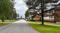 Heikkatie keskellä kuvaa, vasemmalla koivuja ja mäntyjä. Oikealla punatiilirakennuksia. Liikennemerkkit ajokiellosta ja nopeusrajoituksesta. Pilvistä.