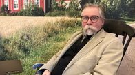 Sulevi Riukulehto istuu keinutuolissa taustallaan lakeusmaisemainen kuvatapetti.