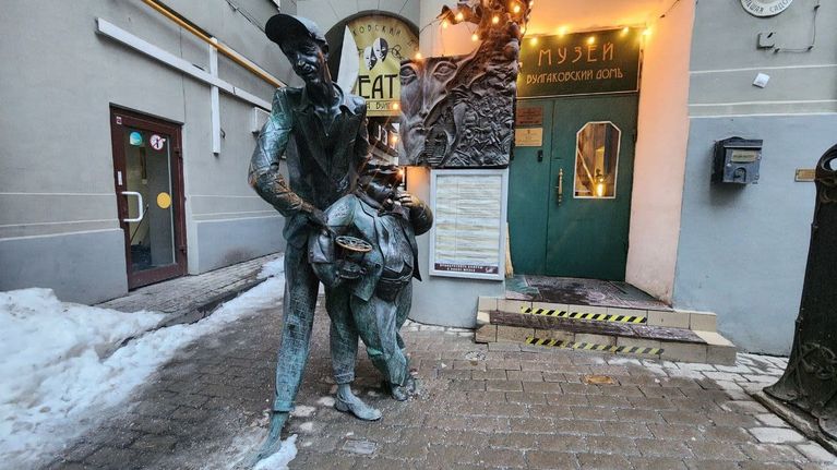 Pitkää laihaa lippalakkipäistä miestä ja lyhyttä lihavaa kissannäköistä miestä esittävä patsas seisoo kerrostalon sisäpihalla vihreän oven vieressä. Oven yllä lukee venäjäksi Museo. Bulgakovin talo.