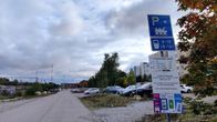 Autoja pysäköitynä hiekkakentälle, jonka vasemmalla puolella tie ja oikealla kerrostaloja. Edessä oikealla pysäköinnistä kertovat kyltit.