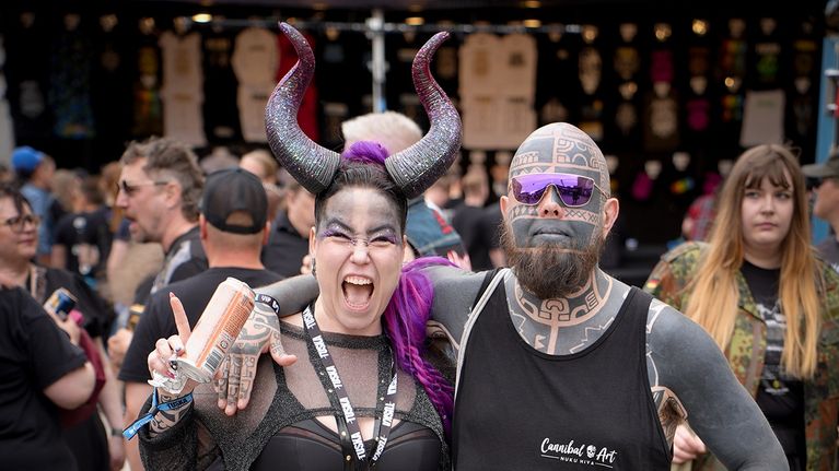 A heavily tattooed man stands with his arm around a woman wearing ram's horns.