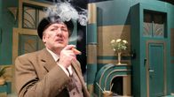 Knallihattupäinen mies polttaa teatterinäyttämöllä sikaria.