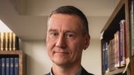 Pukuun pukeutunut mies seisoo kirjahyllyn vieressä.