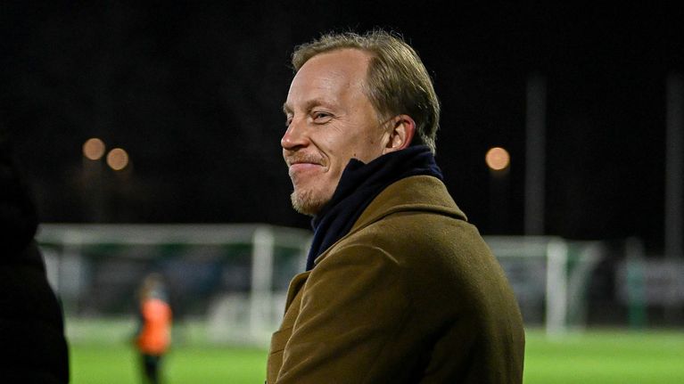 En man i brun rock står vid fotbollsplanen och ler.