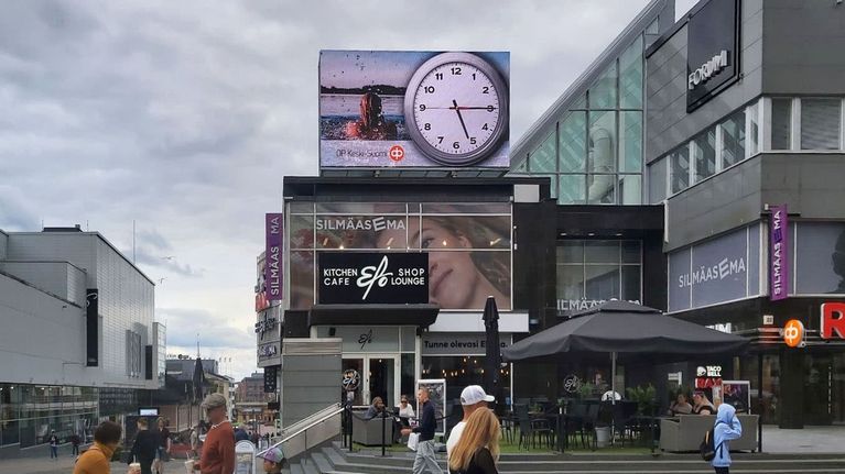 Forumin kauppakeskuksen valotaulussa näkyy Osuuspankin mainos, jossa kello näyttää 5.15. Valotaululn alla on Silmäaseman ja Kitchen Cafe Elosen kyltit. Kauppakeskuksen seinässä lukee Forum. Kadulla kävelee ihmisiä. 