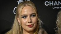 Olivia Oras seisoo mustaa taustaa vasten, jossa näkyy CHANEL- ja muita logoja. Henkilöllä on vaalea tukka ja tummat silmämeikit.