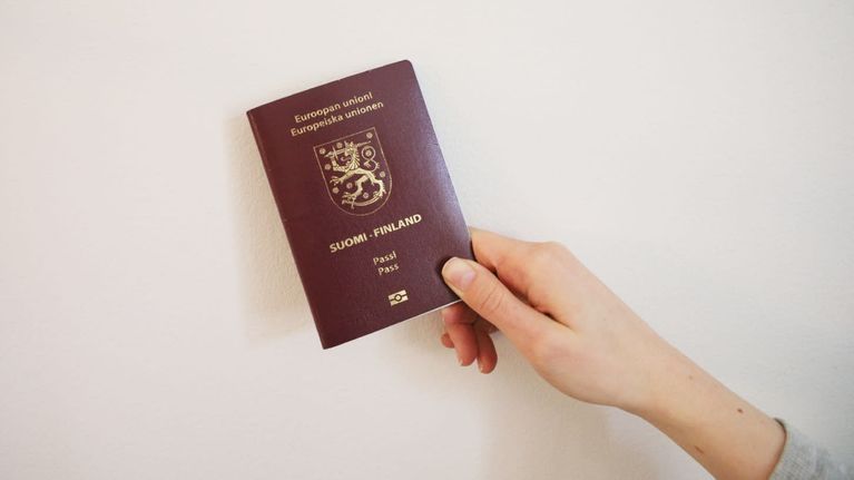 Hand holding a magenta coloured Finnish passport in front of a white wall.