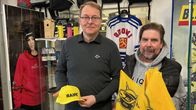 Kaksi henkilöä seisoo jääkiekkotuotteita myyvässä liikkeessä ja pitelevät SaiPa-fanituotteita, kuten lippistä ja keltaisia lippuja.
