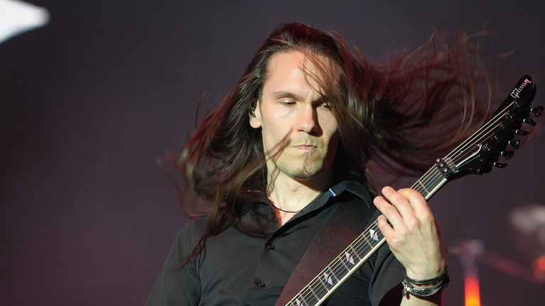 A man with long dark hair, a dark shirt and a serious expression plays electric guitar onstage.