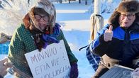 Kyläläisiä pakkasessa osoittamassa mieltä. Huivipäisellä naisella on sylissään kyltti, karvalakkipäinen mies näyttää peukkua.