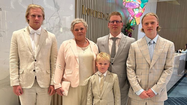 Viisihenkinen perhe seisoo juhlavissa vaaleissa vaatteissa hotellin aulassa ja katsoo hymyillen kameraan.