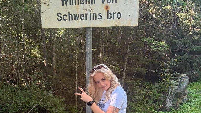 En person står utomhus och pekar mot en skylt med texten "Wilhelm von Schwerins bro" framför skog och grönska.