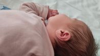 Tummahiuksinen vauva lepää pinnasängyssä vaaleansinisen harson päällä. Kasvot ovat pois päin kamerasta.