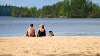Två personer sitter med ryggen mot kameran på en sandstrand. Inte långt från stranden syns en skogsbevuxen holme.