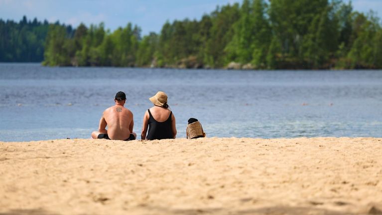 Två personer sitter med ryggen mot kameran på en sandstrand. Inte långt från stranden syns en skogsbevuxen holme.