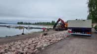 En småbåtshamn med några båtar vid en brygga. På stranden en grävmaskin och två män i neonfärgade skyddsvästar.