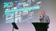 Lyhythiuksinen mies seisoo puhujakorokkeella mikrofoni kädessään, taustalla näkyy esityksen otsikko.