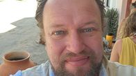 En man med skägg fotograferad utomhus i ett varmt land med träd och hus i bakgrunden.