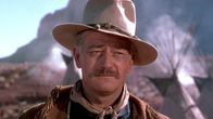 John Wayne hattu päässä, huivi kaulassa ja mokkanahkainen takki harteillaan. Taustalla näkyy intiaanikylä. Kuva elokuvasta Keltainen nauha.