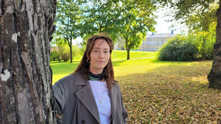 Nainen seisoo puistossa ja nojaa puuhun katsoen kameraa kohti