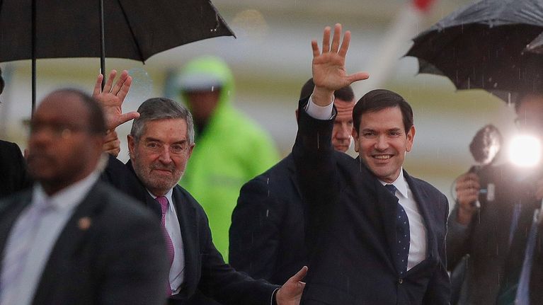 Juan Ramon de la Fuente ja Marco Rubio vilkuttavat ulkona sateessa. Ympärillä ihmisillä sateenvarjoja.