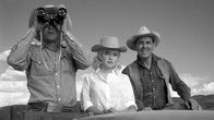 Näyttelijät Montgomery Clift, Marilyn Monroe ja Clark Gable rivissä kuorma-auton lavalla katsovat jonnekin kameran yli etäisyyteen, Cliftillä kiikarit silmillä, miehillä lännenmiehen vaatteet ja hatut, Marilynillä hohtavan valkoinen paita ja hattu; kuva elokuvasta Sopeutumattomat.