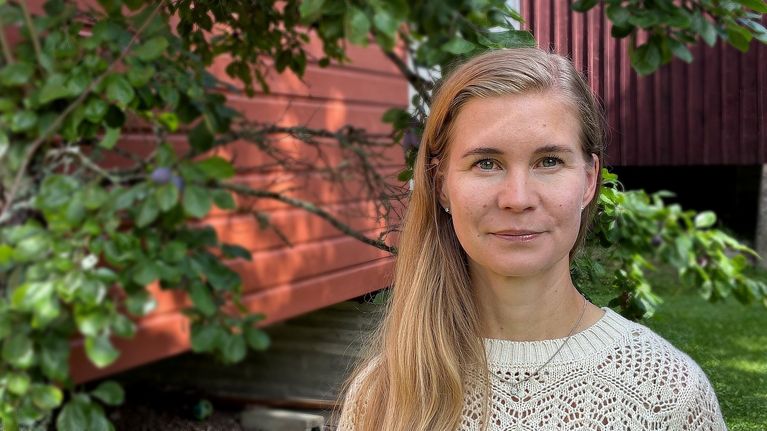 en ung kvinna med husvägg och träd i bakgrunden.