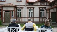 Kuvassa kolme henkilöä istuu ulkona valkoisten pöytien edessä puisia rakennuksia vastapäätä.