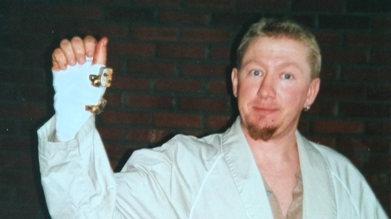 Jari Dahlström i morgonrock står framför en tegelvägg och håller upp en bandagerad hand med ett fingerförband.
