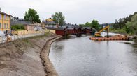 Till vänster Borgå ås östra strand med muren bortplockad och spontar nedslagna i marken. På andra sidan ån står en stor pråm med en orange grävmaskin på.